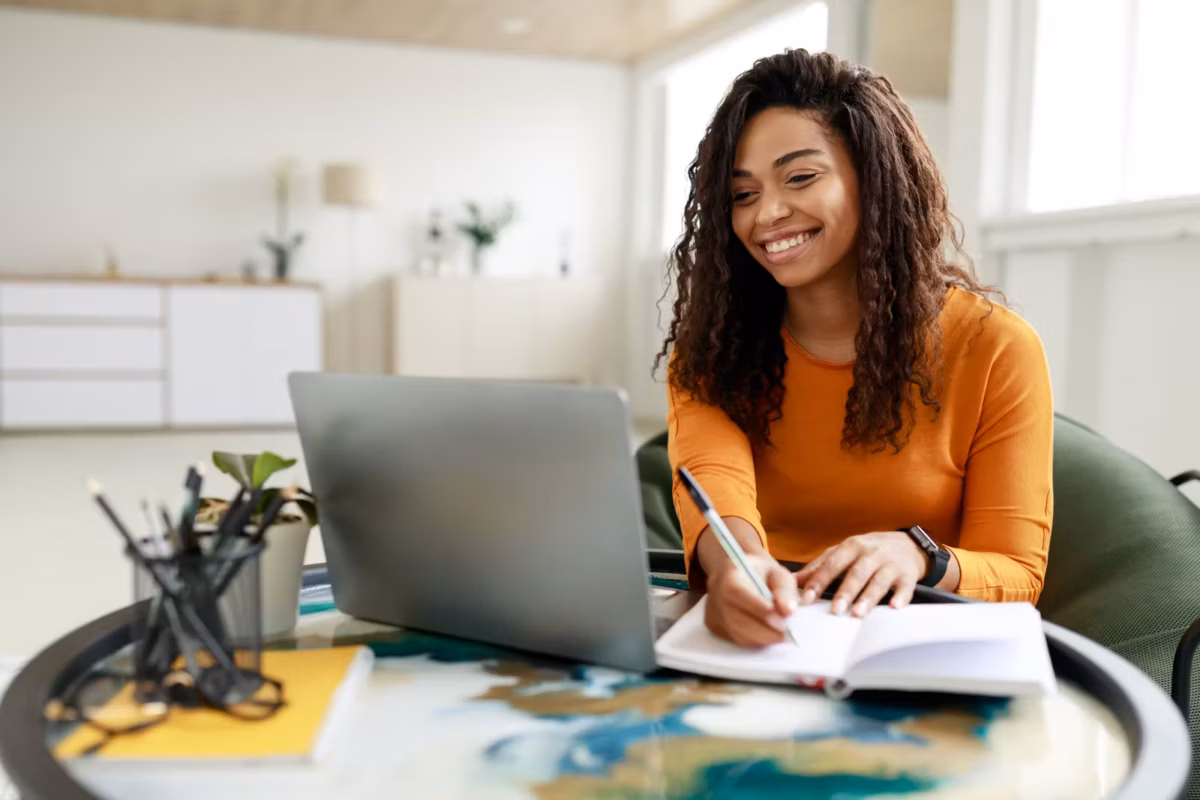 woman writing in notebook and smiling at laptop